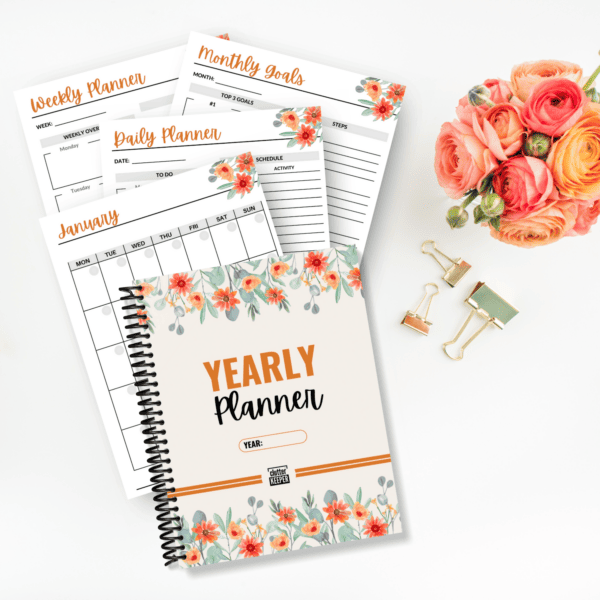 Four example pages and the cover of the Clutter Keeper yearly planner on a desk with a bouquet of orange flowers